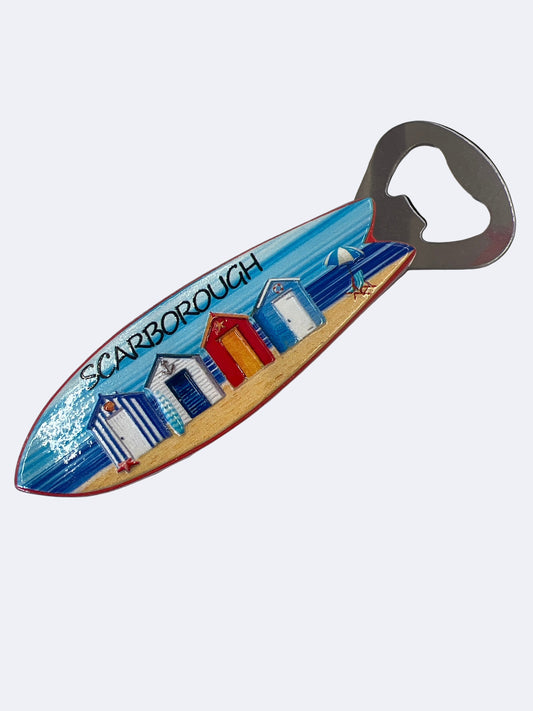 A multicolored surfboard-shaped magnet with a beach and houses design, and a built-in bottle opener.