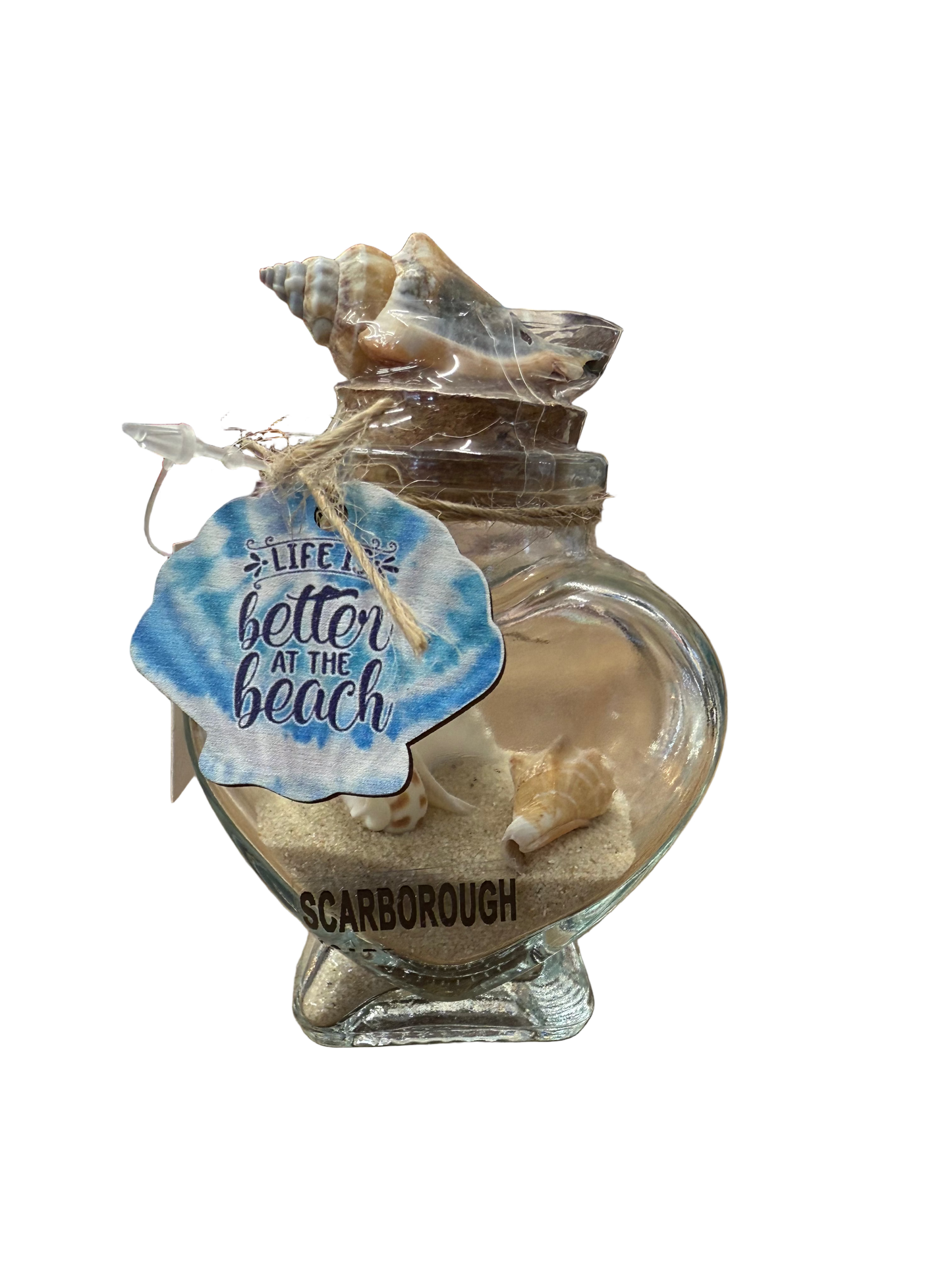 Glass shell-shaped container with sand and a shell, featuring a 'Better at the Beach' tag, on a store counter.
