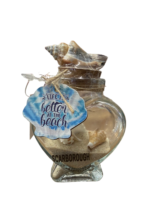 Glass shell-shaped container with sand and a shell, featuring a 'Better at the Beach' tag, on a store counter.